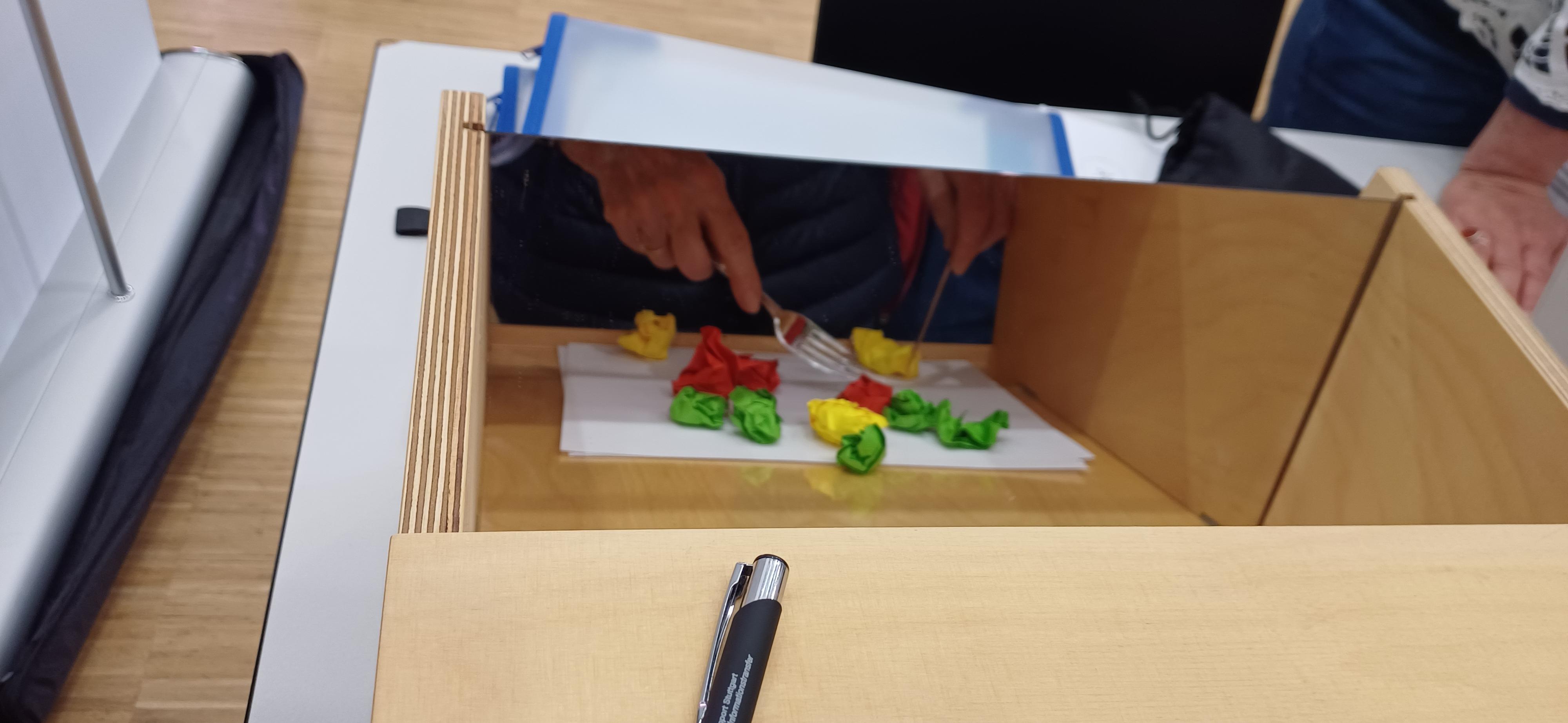 Man sieht einen Spiegel, davor bunte Papierkügelchen, die jemand mit einem Besteck versucht aufzugreifen.